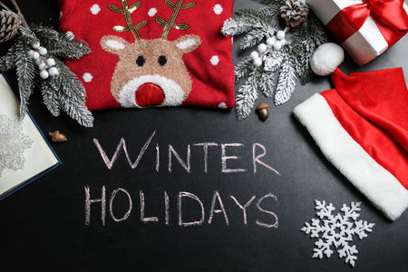 Flat lay composition of Christmas sweater, Santa hat, gift box and decorations on school blackboard with phrase Winter Holidaysの写真素材