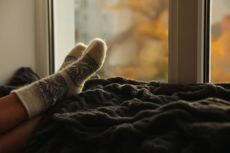 Woman in knitted socks relaxing on plaid near window at home, closeup