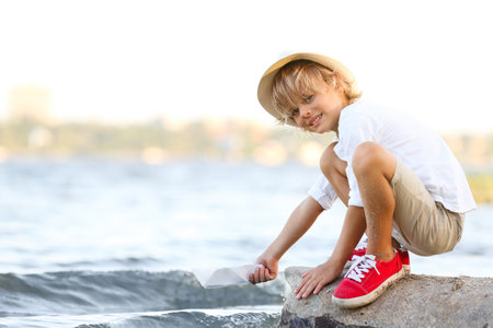Cute little boy with paper ship at river, space for text. child spending time in natureの写真素材