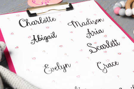 Clipboard with different baby names and toys on gray table, closeup