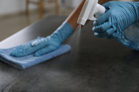 Woman in gloves cleaning gray stone table with rag and detergent indoors, closeup