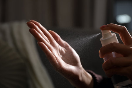 Woman spraying antiseptic onto hand against blurred background, closeup