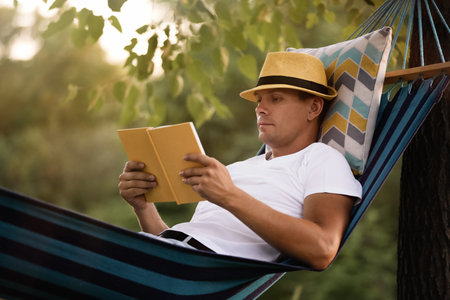 Man reading book in comfortable hammock at green gardenの写真素材