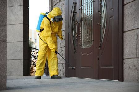 Person in hazmat suit disinfecting entrance door with sprayer. Surface treatment during coronavirus pandemicの写真素材