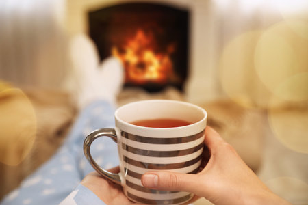Woman with cup of tea resting near fireplace at home, closeupの写真素材