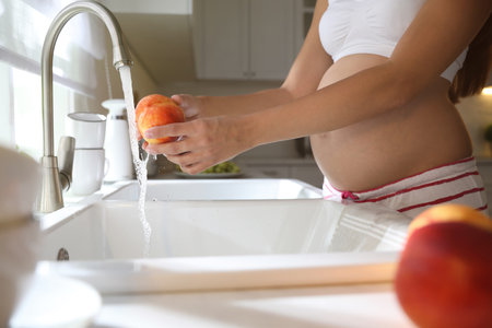 Young pregnant woman washing fresh sweet peach in kitchen, closeup. Taking care of baby healthの写真素材