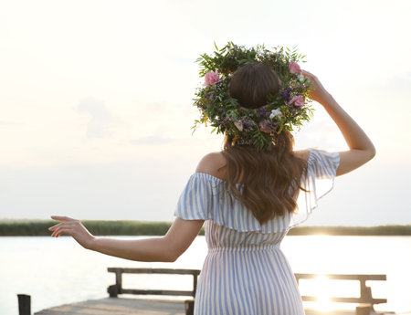 Young woman wearing wreath made of beautiful flowers on pier near river, back viewの写真素材