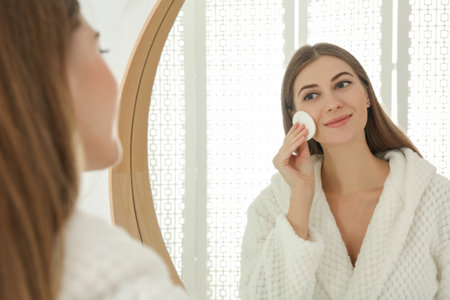 Young woman with cotton pad cleaning her face near mirror in bathroomの写真素材