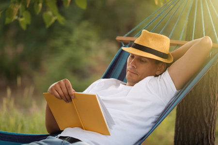 Man reading book in comfortable hammock at green gardenの写真素材