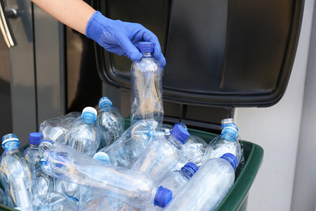 Woman in gloves putting used plastic bottle into trash bin outdoors, closeup. recycling problem
