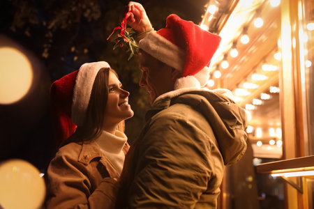 Happy couple in Santa hats standing under mistletoe bunch outdoors, bokeh effectの写真素材