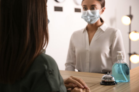 Receptionist with client at countertop in hotel, focus on dispenser bottle of antiseptic gel and service bellの写真素材