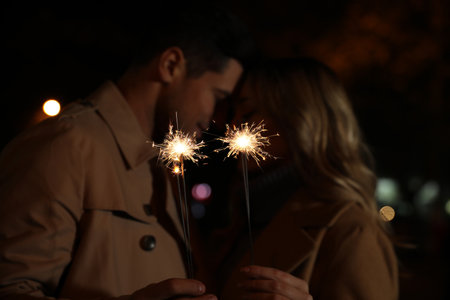 Couple in warm clothes holding burning sparklers at night, focus on fireworks
