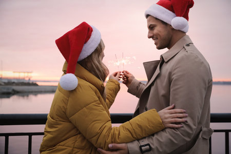 Couple in Santa hats holding burning sparklers near riverの写真素材
