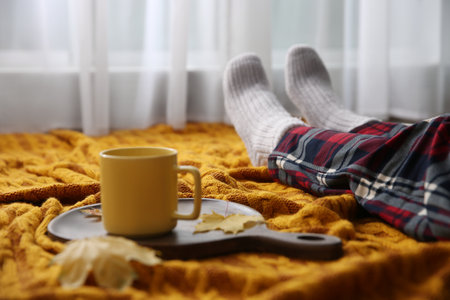 Woman relaxing with cup of hot winter drink on knitted plaid indoors, closeup. Cozy seasonの写真素材