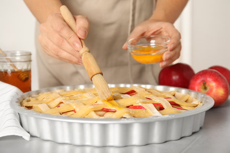 Woman applying liquid egg onto traditional English apple pie with brush at light gray table, closeupの写真素材