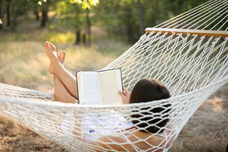 Young woman reading book in comfortable hammock at green gardenの写真素材