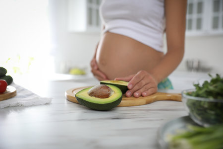 Young pregnant woman with avocado at table in kitchen, closeup. Taking care of baby healthの写真素材