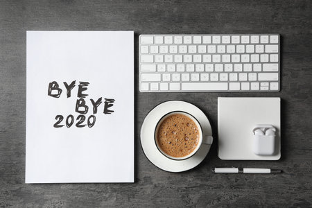 White sheet of paper with phrase Bye Bye 2020 near coffee, keyboard and wireless earphones on gray...