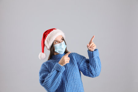 Pretty woman in Santa hat and medical mask pointing on gray backgroundの写真素材