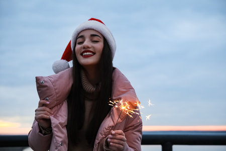 Woman in Santa hat and warm clothes holding burning sparkler outdoorsの写真素材