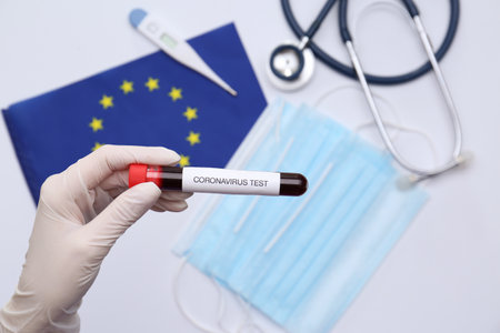 Doctor holding sample tube with label Coronavirus Test above medical items and European Union flag,...
