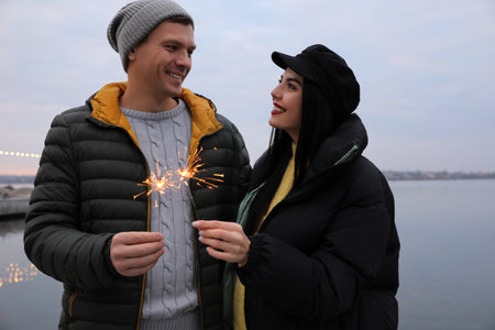 Couple in warm clothes holding burning sparklers near riverの写真素材