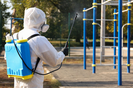 Person in hazmat suit spraying disinfectant around outdoor gym. Surface treatment during coronavirus pandemicの写真素材