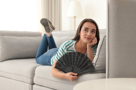 Woman enjoying air flow from hand fan at home. summer heatの写真素材
