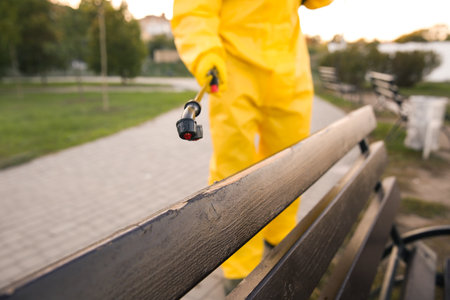 Person in hazmat suit disinfecting bench in park with sprayer, closeup.の写真素材
