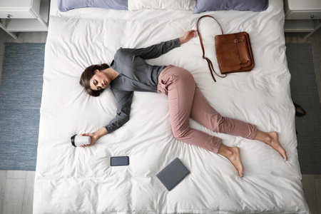 Exhausted woman with cup of coffee sleeping fully dressed on bed at home, above viewの写真素材