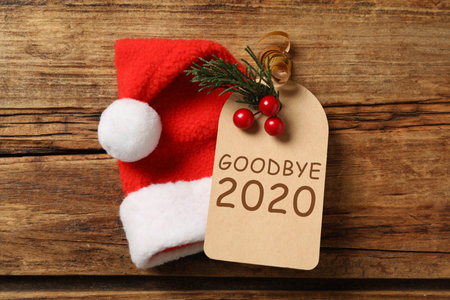 Tag with text Goodbye 2020 and Santa hat on wooden background, flat layの写真素材
