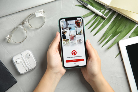 MYKOLAIV, UKRAINE - JULY 9, 2020: Woman holding Iphone 11 with Pinterest app on screen at gray table, top viewのeditorial素材