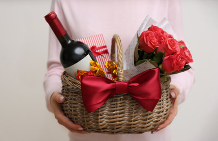 Woman holding wicker basket with gift, bouquet and wine on light gray background, closeupの写真素材