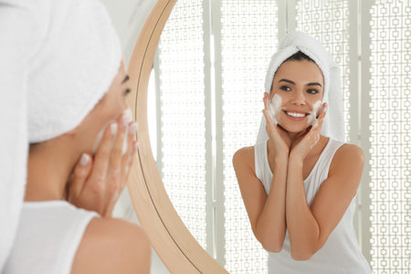 Happy young woman applying cleansing foam onto face near mirror in bathroomの写真素材