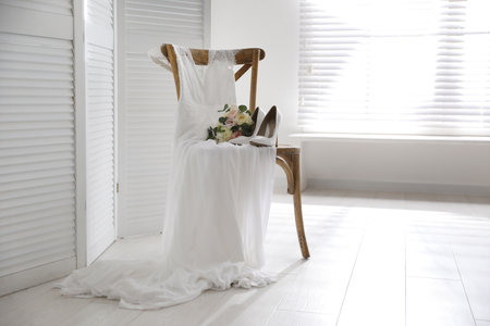 White high heel shoes, flowers and wedding dress on wooden chair indoors. Space for textの写真素材