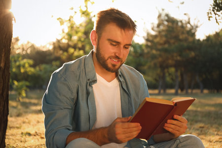 Young man reading book near tree in parkの写真素材