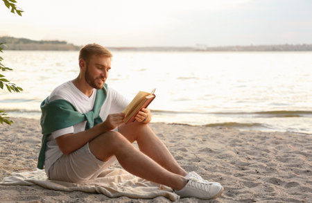 Young man reading book on sandy beach near seaの写真素材