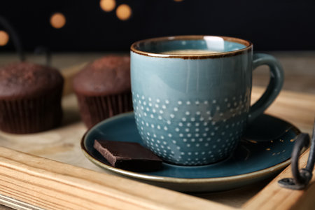 Cup with drink and piece of chocolate on wooden tray against blurred lights, closeupの写真素材