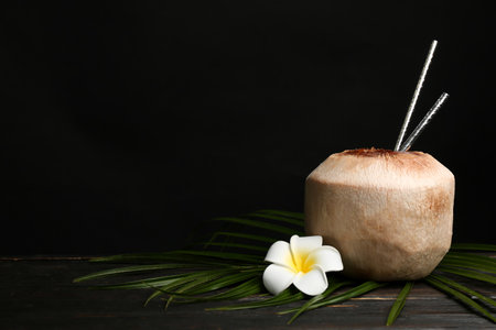 Fresh coconut with drinking straws and flower on wooden table against black background. Space for textの写真素材