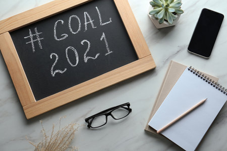 Chalkboard with hashtag Goal 2021 near smartphone and notebooks on white table, flat lay. New year targetsの写真素材