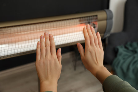 Woman warming hands near electric infrared heater indoors, closeupの写真素材