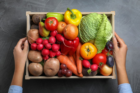 Farmer with wooden crate full of different vegetables and fruits at grey table, top view. Harvesting timeの写真素材