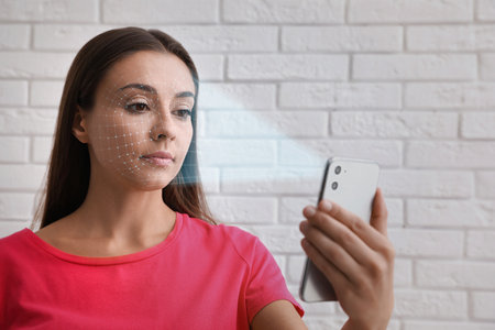 Young woman unlocking smartphone with facial scanner near white brick wall. biometric verificationの写真素材
