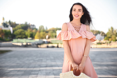 Beautiful young woman in stylish pink dress with handbag on city streetの写真素材