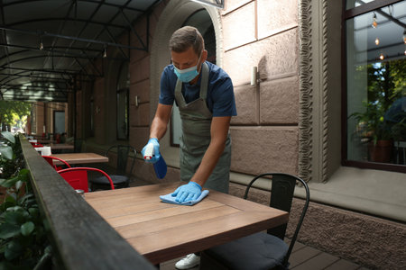 Waiter in mask and gloves disinfecting table at outdoor cafeの写真素材