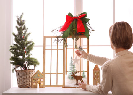 Woman creating Christmas composition inside of vintage wooden lantern on window sill indoorsの写真素材