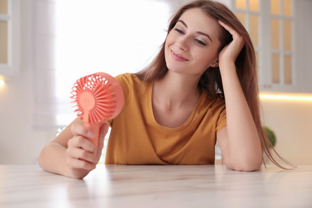 Woman enjoying air flow from portable fan at table in kitchen. summer heatの写真素材