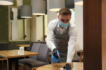 Waiter cleaning table with rag and detergent in restaurant. Surface treatment during coronavirus quarantineの写真素材
