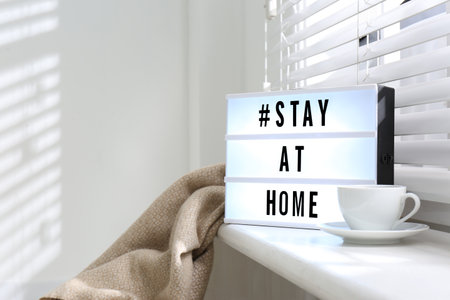 Cup of drink and lightbox with hashtag STAY AT HOME on window sill indoors. Message to promote self-isolation during COVID?19 pandemicの写真素材